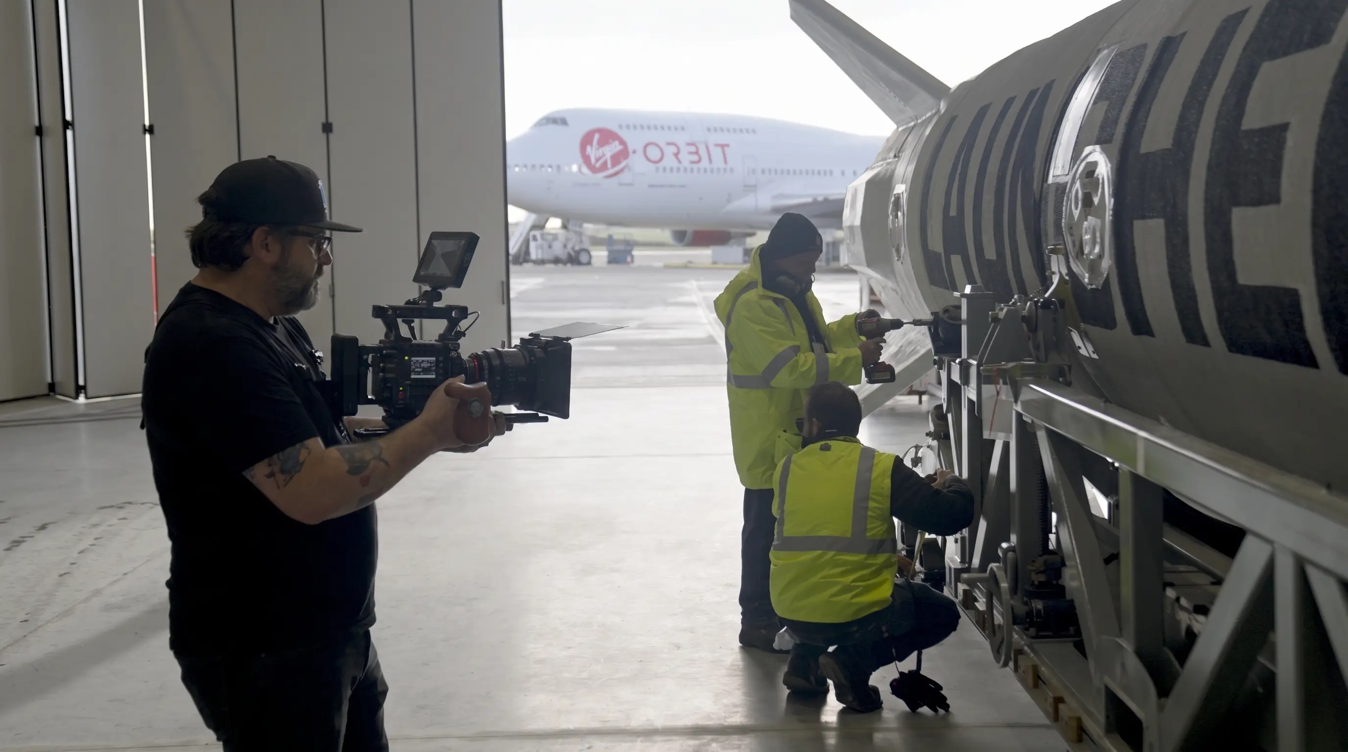Daniel Jarvis filming technicians working on LauncherOne, with Virgin Orbit’s Cosmic Girl aircraft outside the hangar.
